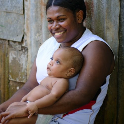 A mother holds her child in her arms while smiling and sitting against the wall of a corrugated steel building.