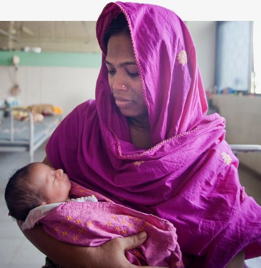 A mother wearing a pink hijab holds her infant child in her arms in a hospital room.