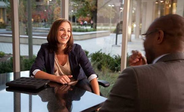 Woman sitting at conference table in front of large windows in urban setting, smiling at man across the table, in an interview or meeting context