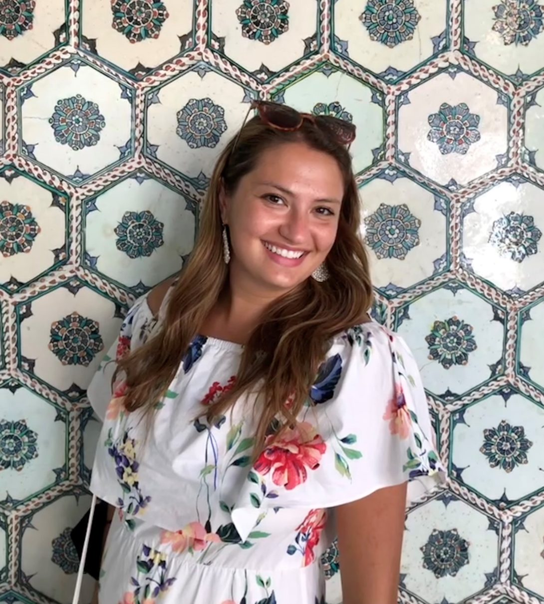 Portrait of woman with long light brown hair and sunglasses on top of head smiling at camera at a slight angle in front of tiled patterned wall