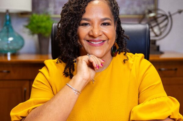 Woman in bright yellow shirt smiling at camera with hand under chin and elbows resting on table