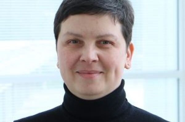 Person in black turtleneck with very short dark hair and light blue window-like background, smiling without teeth at camera