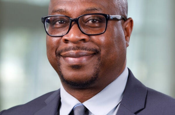 Black bald man with dark rimmed glasses and mustache in darker suit with light blue button up and striped tie smiling without teeth at camera