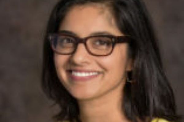 Woman with shoulder length dark hair and dark-rimmed glasses in yellow shirt, smiling at camera