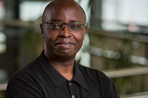 Black man with bald head and glasses in dark button up shirt with crossed arms, smiling closed-mouth at camera