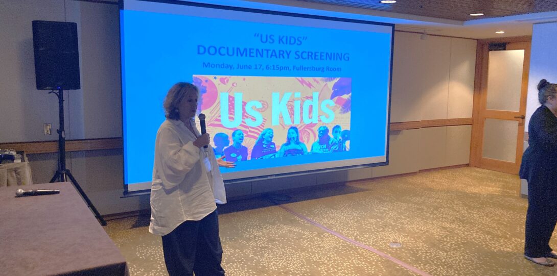 Person with mic standing in front of screen that says Us Kids