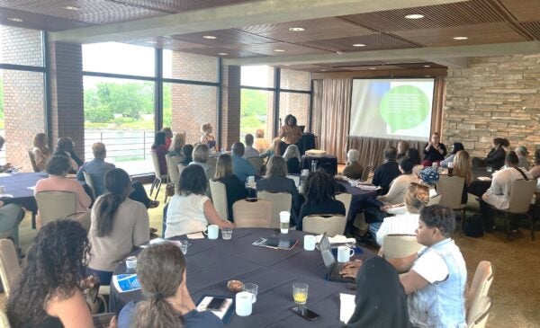 Room full of people at round tables with screen in center back of photo and speaker standing in front of screen