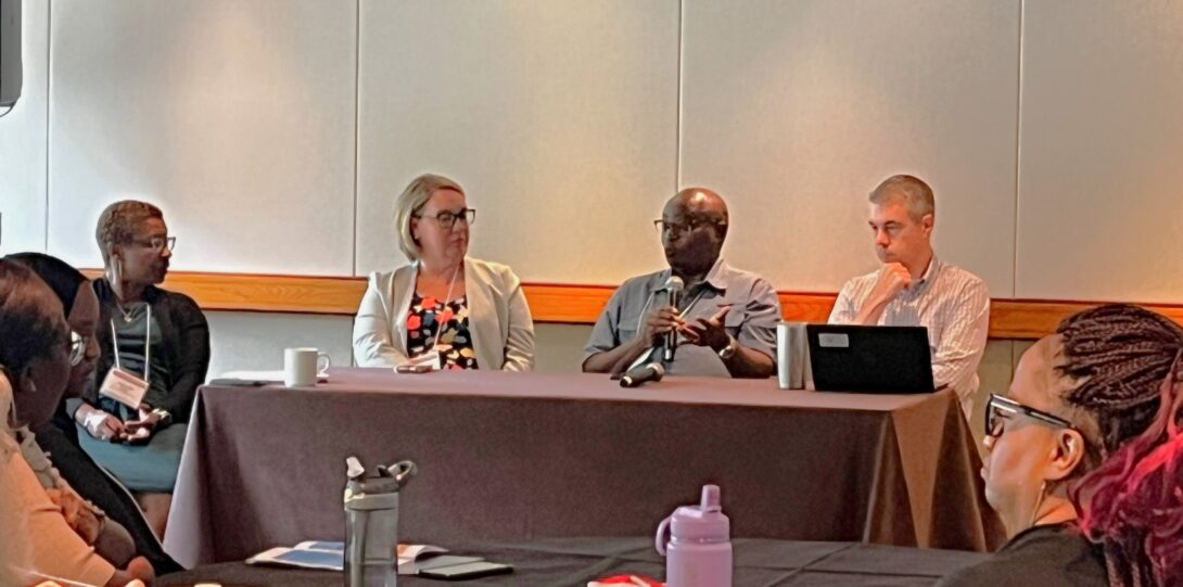 three people sitting at table in center of room facing camera with person moderating panel on left of image