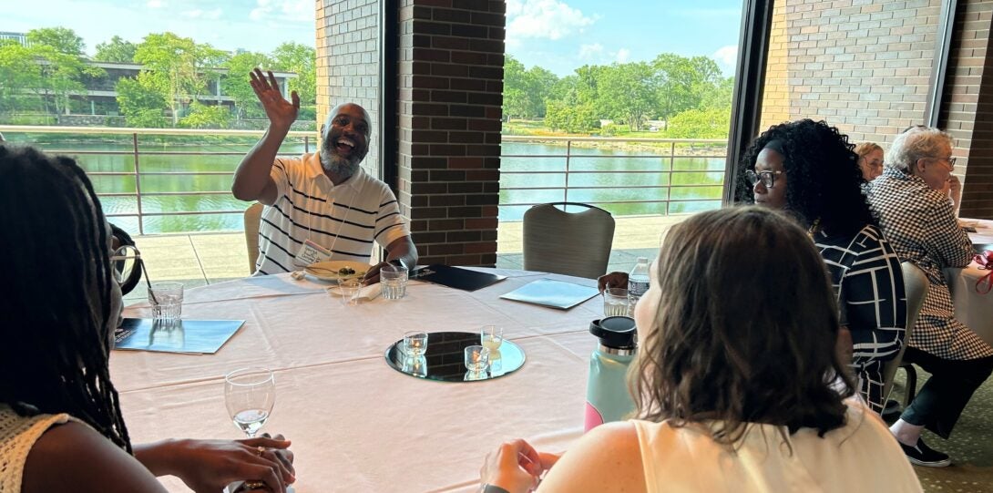 table of people in front of windows looking out at lake and blue sky, with person facing camera in center of shot waving and smiling at camera