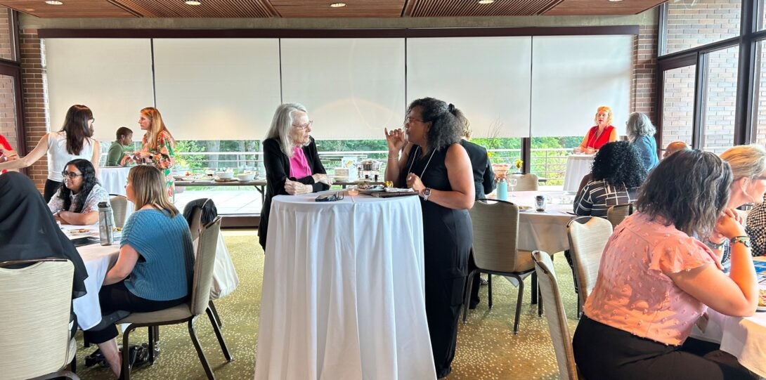 two people standing at tallboy table talking surrounded by others sitting at round tables