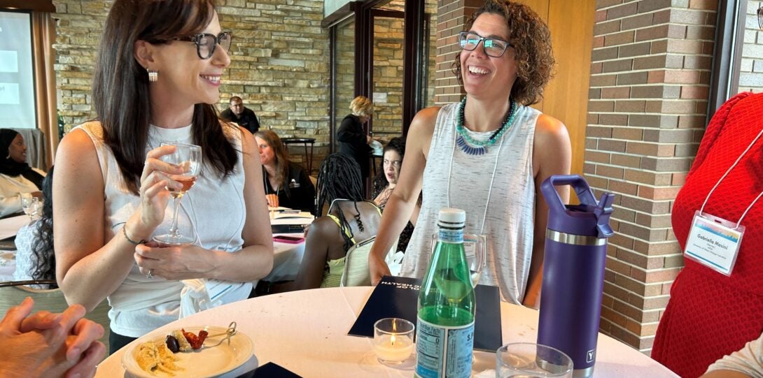 Two women wearing glasses with one holding a wine glass standing at tallboy table smiling and laughing